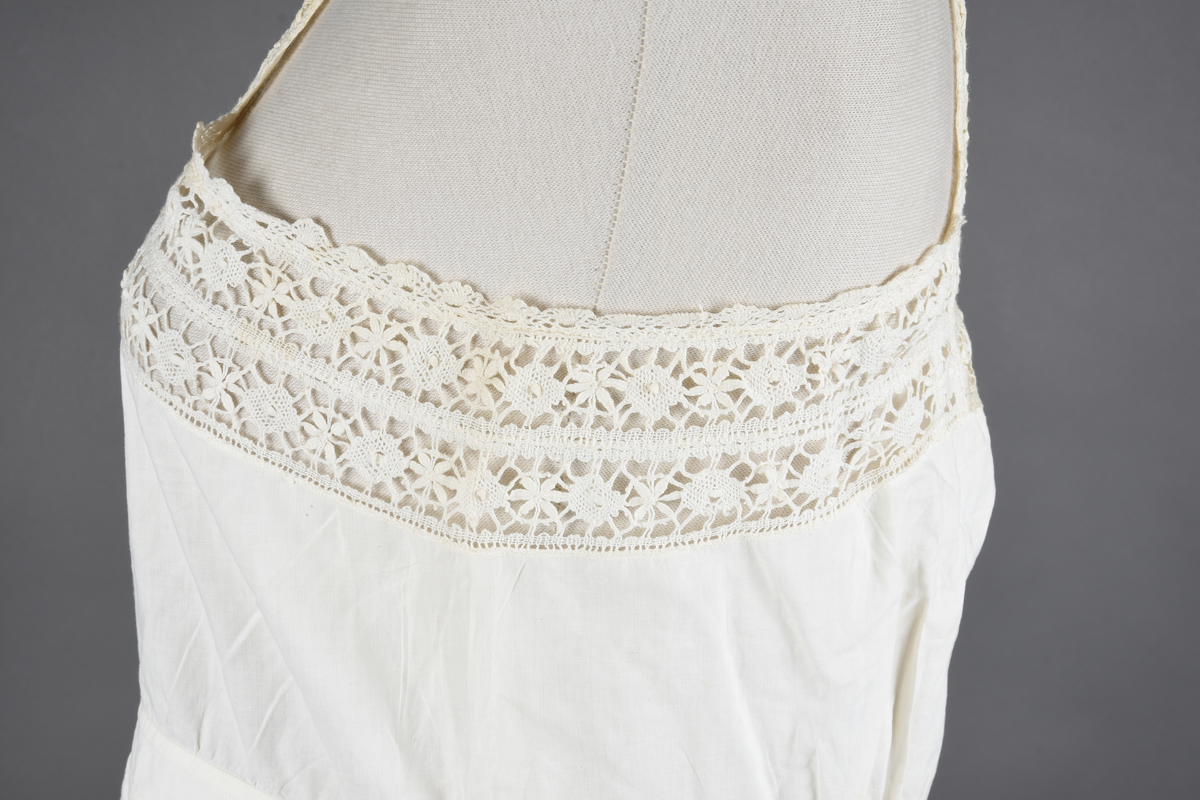 Kombinasjon av fint, hvitt bomullslerret. Livet har smale blonde-skulderstropper festet mot en bred blondekant. Foran er kombinasjonen rynket til en linning i taillen. Benklærne er sydd til samme linning og de har lavt skritt og er helt åpne nedentil. Nederst er de kantet med rynkekappe og blonder. Knappet foran med ti små knapper. Noen rustflekker. Lite hull revet bak.