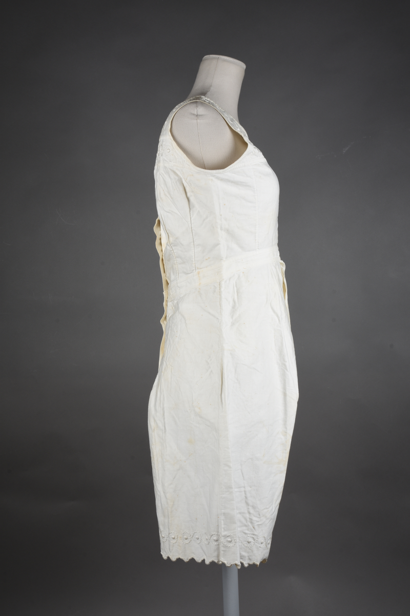 Kombinasjon av bomullslerret. Kraftig kypervevd lerret (twill). Overdelen har brede skulderstropper og er figursydd til en linning i taillen. Benklærne er sydd til samme linning og de har lavt skritt og er helt åpne nedentil. Knappet foran med elleve små knapper. Broderi og tungekant nederst - “løpende hund”-ranke rundt store prikker, tungekant på beina. Noen rustflekker. Lite hull revet bak.