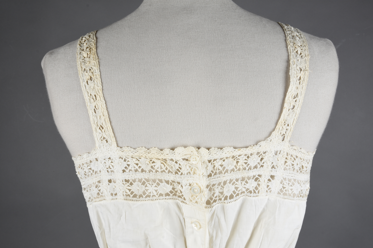 Kombinasjon av fint, hvitt bomullslerret. Livet har smale blonde-skulderstropper festet mot en bred blondekant. Foran er kombinasjonen rynket til en linning i taillen. Benklærne er sydd til samme linning og de har lavt skritt og er helt åpne nedentil. Nederst er de kantet med rynkekappe og blonder. Knappet foran med ti små knapper. Noen rustflekker. Lite hull revet bak.