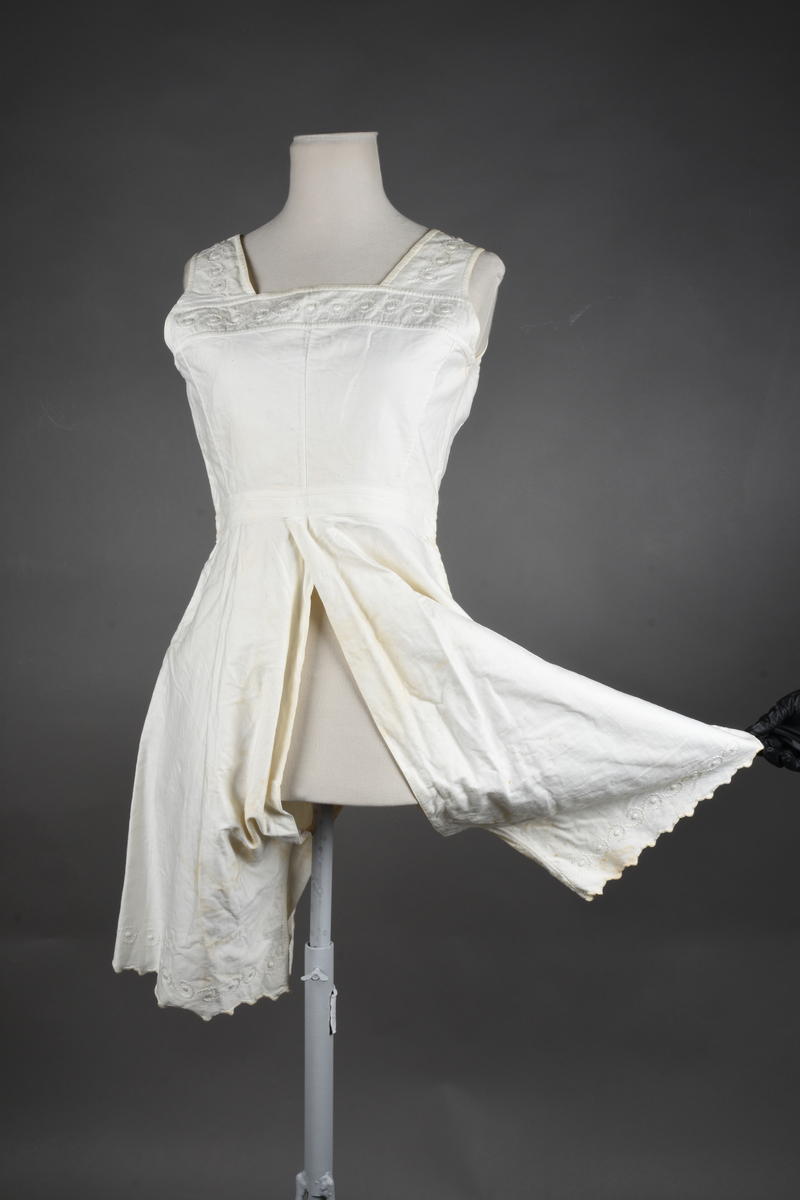 Kombinasjon av bomullslerret. Kraftig kypervevd lerret (twill). Overdelen har brede skulderstropper og er figursydd til en linning i taillen. Benklærne er sydd til samme linning og de har lavt skritt og er helt åpne nedentil. Knappet foran med elleve små knapper. Broderi og tungekant nederst - “løpende hund”-ranke rundt store prikker, tungekant på beina. Noen rustflekker. Lite hull revet bak.