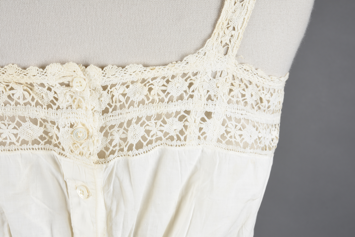 Kombinasjon av fint, hvitt bomullslerret. Livet har smale blonde-skulderstropper festet mot en bred blondekant. Foran er kombinasjonen rynket til en linning i taillen. Benklærne er sydd til samme linning og de har lavt skritt og er helt åpne nedentil. Nederst er de kantet med rynkekappe og blonder. Knappet foran med ti små knapper. Noen rustflekker. Lite hull revet bak.