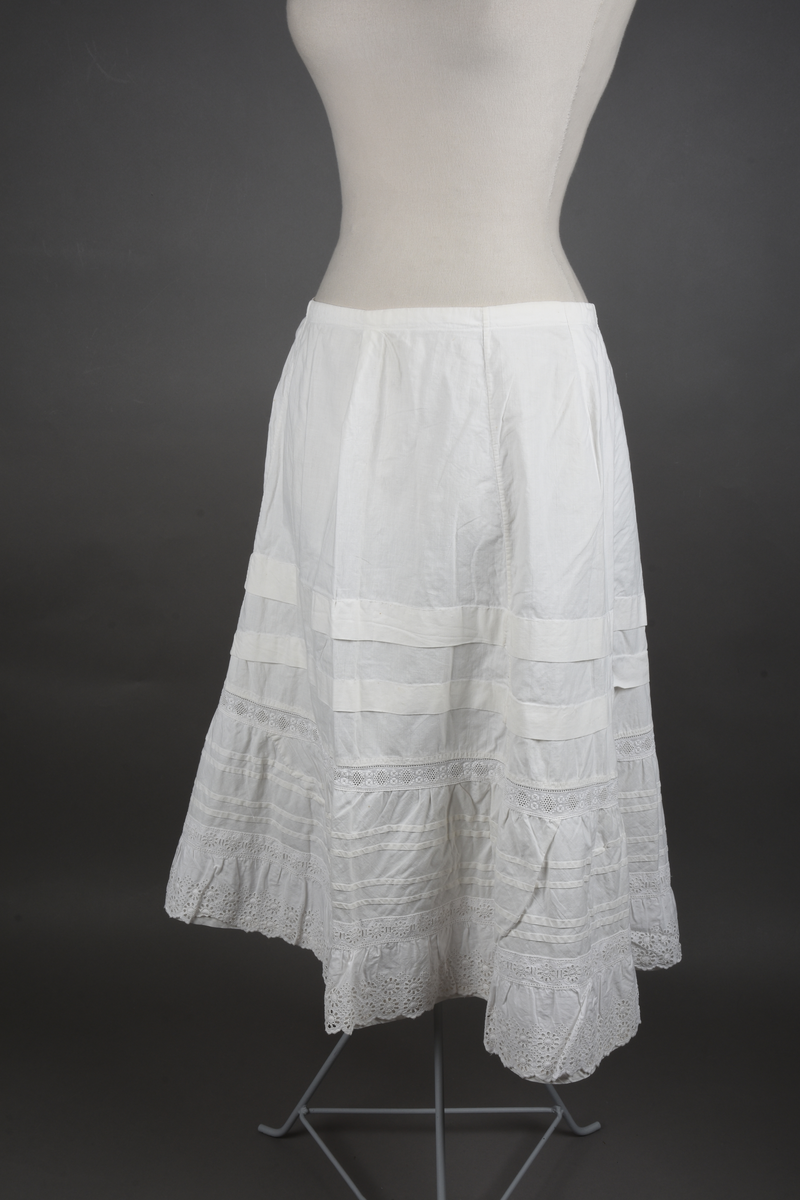 Underskjørt av lin. Sveitserblonde av bomull. Skjørtet er sydd i fem bredder, hver med et innsnitt oppe. En løpegang i øvre kant tyder på at bindebåndet opprinnelig har vært et trekkebånd, men nå er det bare sydd fast i hver side av splitten. Nede er det en 26 cm bred kappe, skåret på skrå av stoffet og lett rynket til et mellomverk i overgangen til selve skjørtet. Kappen er stivet av med fire bieser og dessuten blitt påsydd (utenpå, ikke nedenfor) et mellomverk og en rynkekappe av sveitserblonde. Ovenfor kappene er skjørtet forkortet av to tråklete legg, hver ca 3 cm brede.