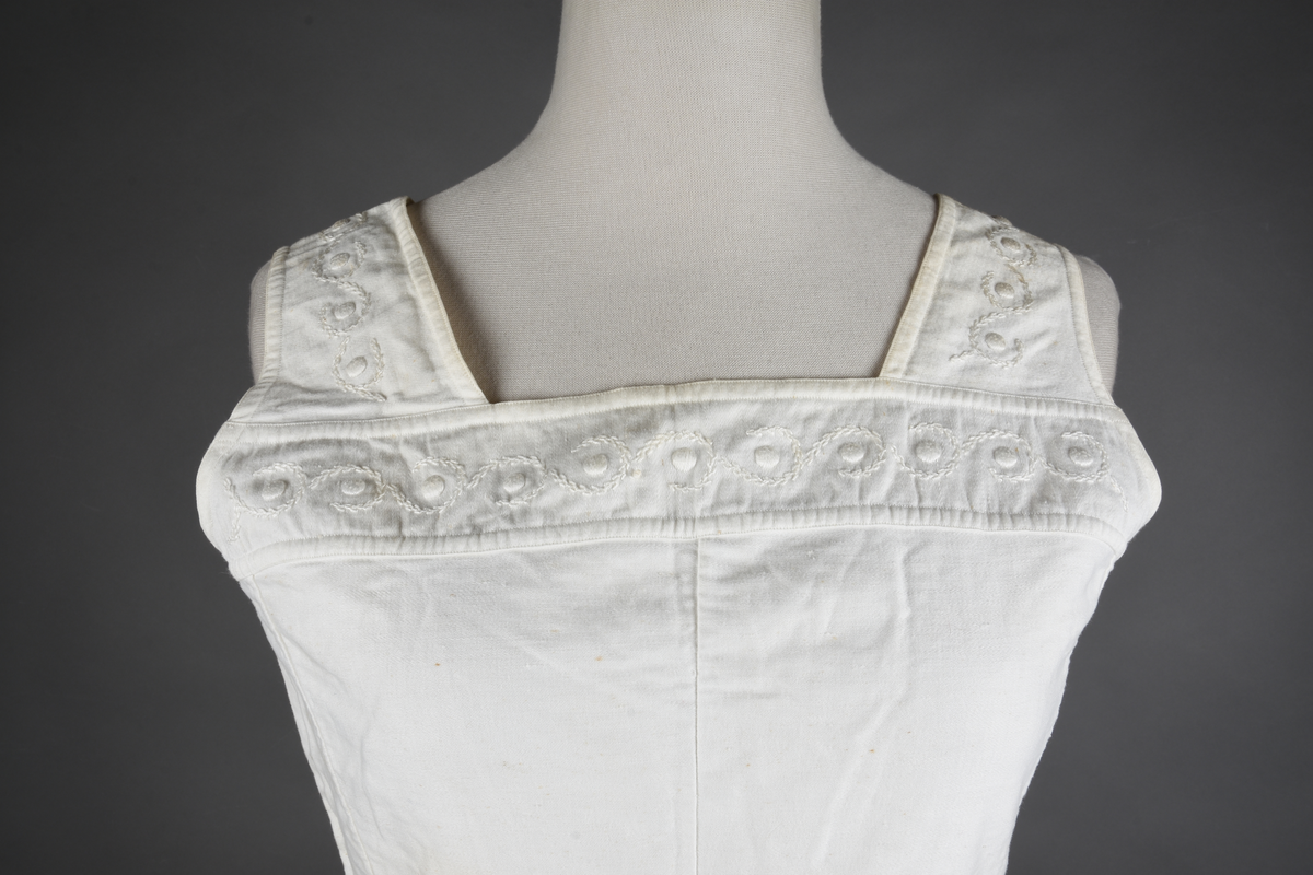 Kombinasjon av bomullslerret. Kraftig kypervevd lerret (twill). Overdelen har brede skulderstropper og er figursydd til en linning i taillen. Benklærne er sydd til samme linning og de har lavt skritt og er helt åpne nedentil. Knappet foran med elleve små knapper. Broderi og tungekant nederst - “løpende hund”-ranke rundt store prikker, tungekant på beina. Noen rustflekker. Lite hull revet bak.