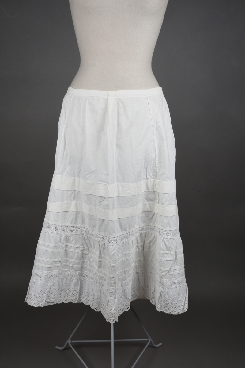 Underskjørt av lin. Sveitserblonde av bomull. Skjørtet er sydd i fem bredder, hver med et innsnitt oppe. En løpegang i øvre kant tyder på at bindebåndet opprinnelig har vært et trekkebånd, men nå er det bare sydd fast i hver side av splitten. Nede er det en 26 cm bred kappe, skåret på skrå av stoffet og lett rynket til et mellomverk i overgangen til selve skjørtet. Kappen er stivet av med fire bieser og dessuten blitt påsydd (utenpå, ikke nedenfor) et mellomverk og en rynkekappe av sveitserblonde. Ovenfor kappene er skjørtet forkortet av to tråklete legg, hver ca 3 cm brede.