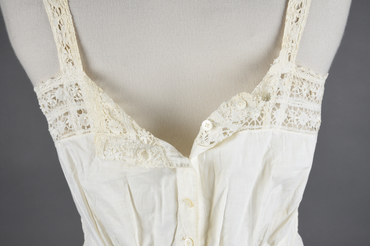 Kombinasjon av fint, hvitt bomullslerret. Livet har smale blonde-skulderstropper festet mot en bred blondekant. Foran er kombinasjonen rynket til en linning i taillen. Benklærne er sydd til samme linning og de har lavt skritt og er helt åpne nedentil. Nederst er de kantet med rynkekappe og blonder. Knappet foran med ti små knapper. Noen rustflekker. Lite hull revet bak.