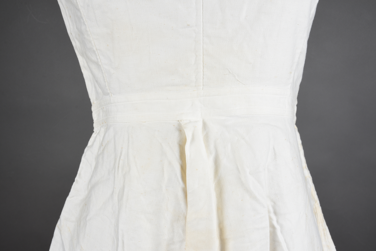 Kombinasjon av bomullslerret. Kraftig kypervevd lerret (twill). Overdelen har brede skulderstropper og er figursydd til en linning i taillen. Benklærne er sydd til samme linning og de har lavt skritt og er helt åpne nedentil. Knappet foran med elleve små knapper. Broderi og tungekant nederst - “løpende hund”-ranke rundt store prikker, tungekant på beina. Noen rustflekker. Lite hull revet bak.