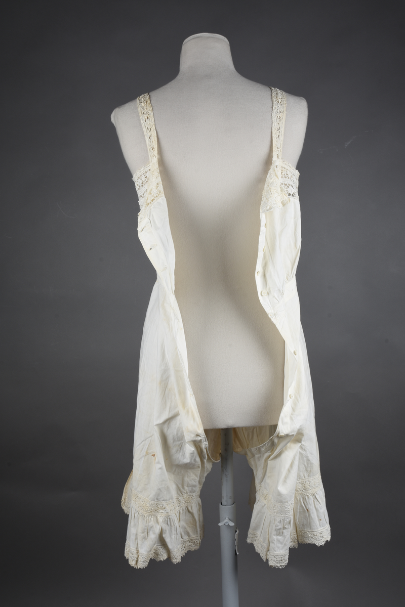 Kombinasjon av fint, hvitt bomullslerret. Livet har smale blonde-skulderstropper festet mot en bred blondekant. Foran er kombinasjonen rynket til en linning i taillen. Benklærne er sydd til samme linning og de har lavt skritt og er helt åpne nedentil. Nederst er de kantet med rynkekappe og blonder. Knappet foran med ti små knapper. Noen rustflekker. Lite hull revet bak.