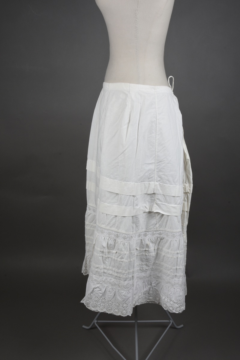 Underskjørt av lin. Sveitserblonde av bomull. Skjørtet er sydd i fem bredder, hver med et innsnitt oppe. En løpegang i øvre kant tyder på at bindebåndet opprinnelig har vært et trekkebånd, men nå er det bare sydd fast i hver side av splitten. Nede er det en 26 cm bred kappe, skåret på skrå av stoffet og lett rynket til et mellomverk i overgangen til selve skjørtet. Kappen er stivet av med fire bieser og dessuten blitt påsydd (utenpå, ikke nedenfor) et mellomverk og en rynkekappe av sveitserblonde. Ovenfor kappene er skjørtet forkortet av to tråklete legg, hver ca 3 cm brede.