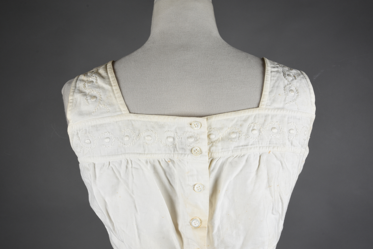 Kombinasjon av bomullslerret. Kraftig kypervevd lerret (twill). Overdelen har brede skulderstropper og er figursydd til en linning i taillen. Benklærne er sydd til samme linning og de har lavt skritt og er helt åpne nedentil. Knappet foran med elleve små knapper. Broderi og tungekant nederst - “løpende hund”-ranke rundt store prikker, tungekant på beina. Noen rustflekker. Lite hull revet bak.