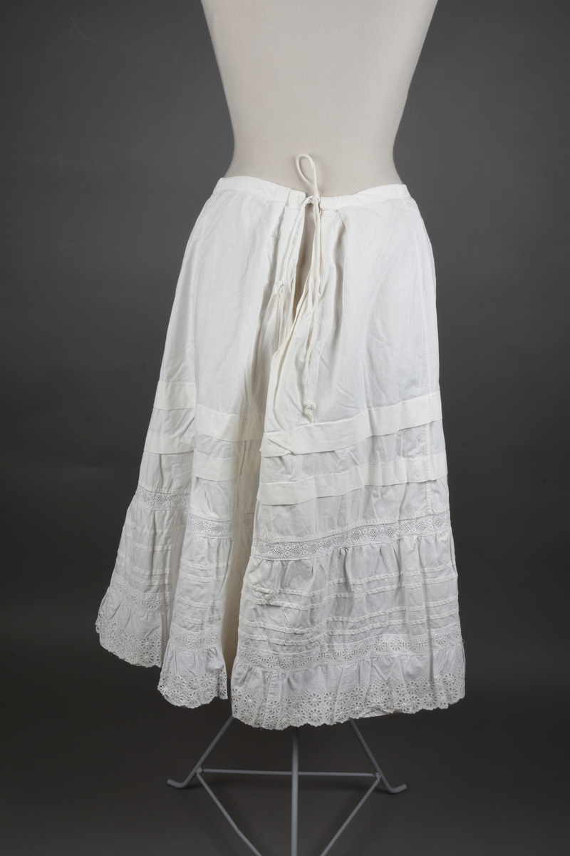 Underskjørt av lin. Sveitserblonde av bomull. Skjørtet er sydd i fem bredder, hver med et innsnitt oppe. En løpegang i øvre kant tyder på at bindebåndet opprinnelig har vært et trekkebånd, men nå er det bare sydd fast i hver side av splitten. Nede er det en 26 cm bred kappe, skåret på skrå av stoffet og lett rynket til et mellomverk i overgangen til selve skjørtet. Kappen er stivet av med fire bieser og dessuten blitt påsydd (utenpå, ikke nedenfor) et mellomverk og en rynkekappe av sveitserblonde. Ovenfor kappene er skjørtet forkortet av to tråklete legg, hver ca 3 cm brede.