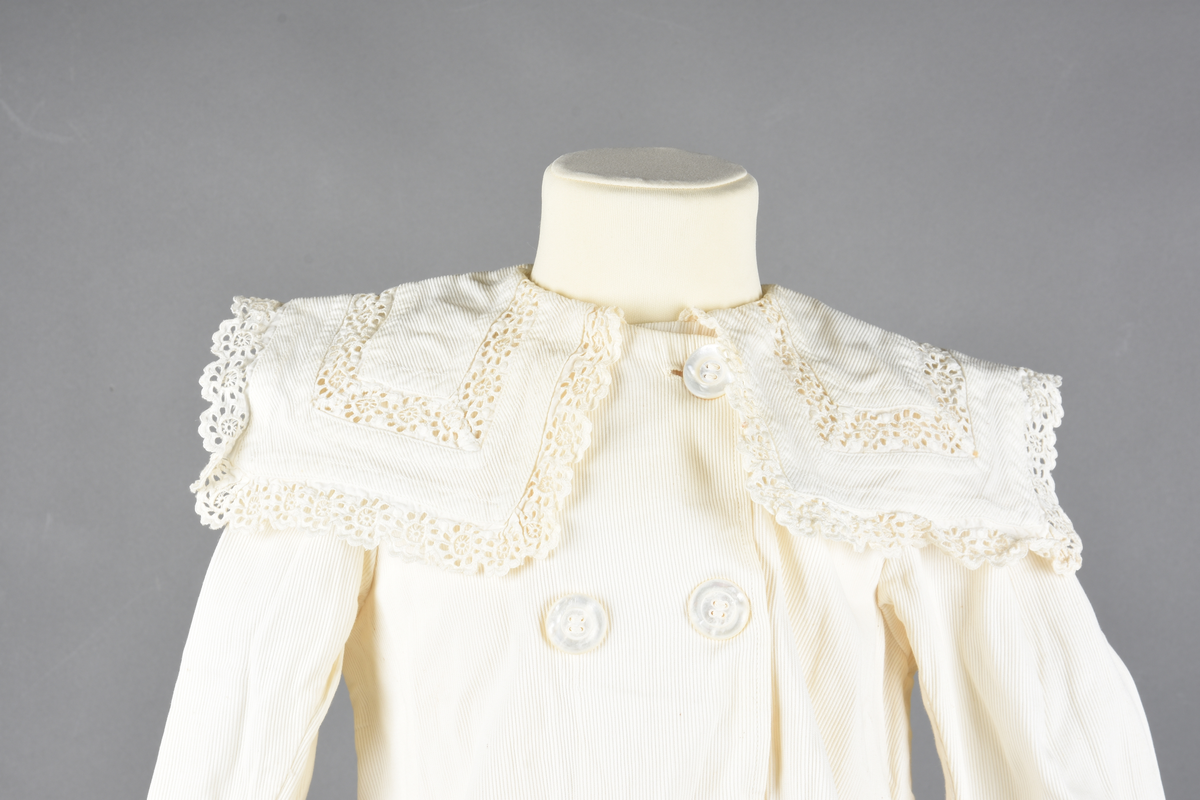 Jakkke av bomull. Dobbelspent med seks knapper. Stor firkantet “matros”- krage med mellomverk og kant av sveitserblonde. Opphengs-løkker sydd inn i ermegapene.
