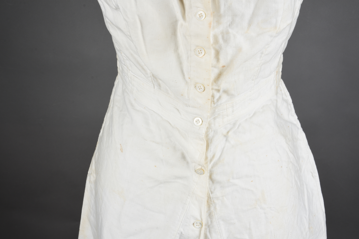 Kombinasjon av bomullslerret. Kraftig kypervevd lerret (twill). Overdelen har brede skulderstropper og er figursydd til en linning i taillen. Benklærne er sydd til samme linning og de har lavt skritt og er helt åpne nedentil. Knappet foran med elleve små knapper. Broderi og tungekant nederst - “løpende hund”-ranke rundt store prikker, tungekant på beina. Noen rustflekker. Lite hull revet bak.