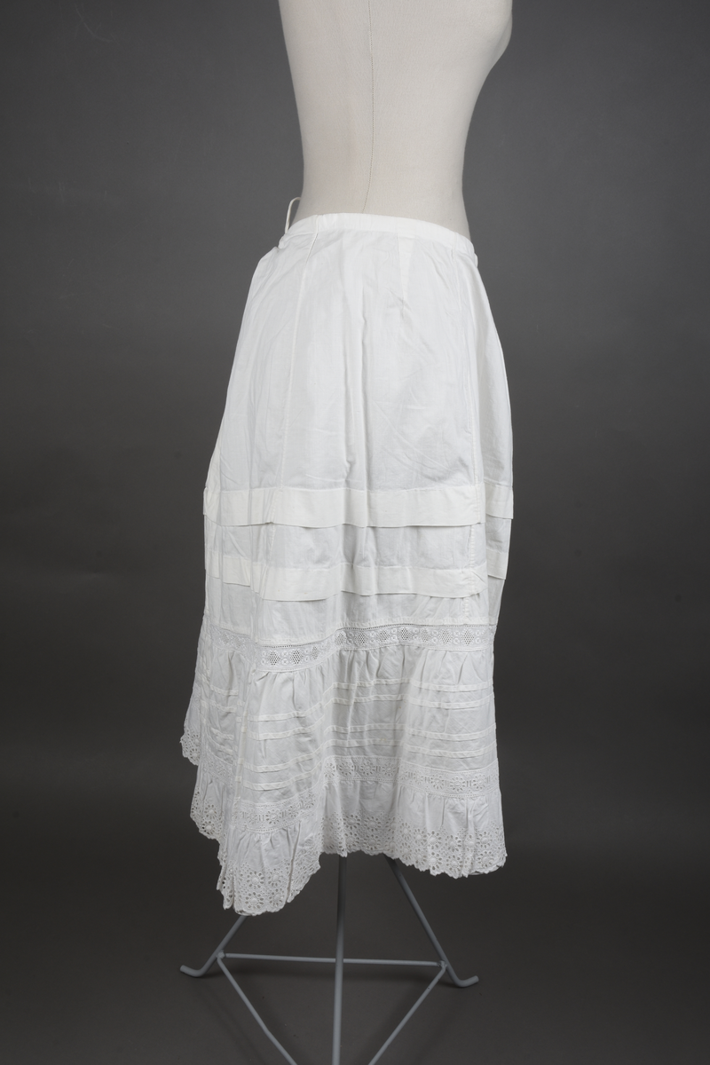 Underskjørt av lin. Sveitserblonde av bomull. Skjørtet er sydd i fem bredder, hver med et innsnitt oppe. En løpegang i øvre kant tyder på at bindebåndet opprinnelig har vært et trekkebånd, men nå er det bare sydd fast i hver side av splitten. Nede er det en 26 cm bred kappe, skåret på skrå av stoffet og lett rynket til et mellomverk i overgangen til selve skjørtet. Kappen er stivet av med fire bieser og dessuten blitt påsydd (utenpå, ikke nedenfor) et mellomverk og en rynkekappe av sveitserblonde. Ovenfor kappene er skjørtet forkortet av to tråklete legg, hver ca 3 cm brede.