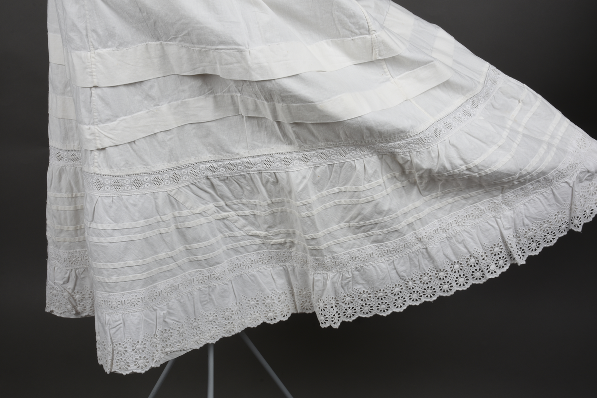 Underskjørt av lin. Sveitserblonde av bomull. Skjørtet er sydd i fem bredder, hver med et innsnitt oppe. En løpegang i øvre kant tyder på at bindebåndet opprinnelig har vært et trekkebånd, men nå er det bare sydd fast i hver side av splitten. Nede er det en 26 cm bred kappe, skåret på skrå av stoffet og lett rynket til et mellomverk i overgangen til selve skjørtet. Kappen er stivet av med fire bieser og dessuten blitt påsydd (utenpå, ikke nedenfor) et mellomverk og en rynkekappe av sveitserblonde. Ovenfor kappene er skjørtet forkortet av to tråklete legg, hver ca 3 cm brede.