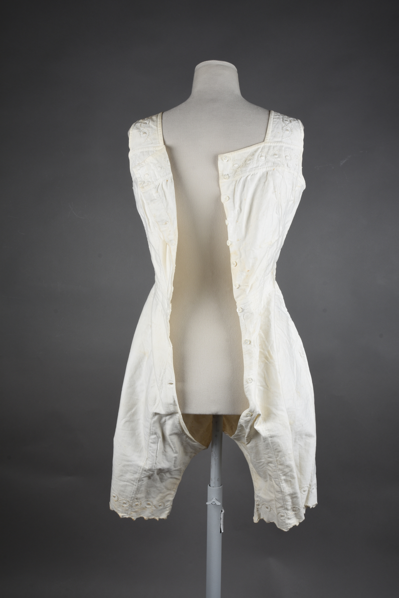 Kombinasjon av bomullslerret. Kraftig kypervevd lerret (twill). Overdelen har brede skulderstropper og er figursydd til en linning i taillen. Benklærne er sydd til samme linning og de har lavt skritt og er helt åpne nedentil. Knappet foran med elleve små knapper. Broderi og tungekant nederst - “løpende hund”-ranke rundt store prikker, tungekant på beina. Noen rustflekker. Lite hull revet bak.