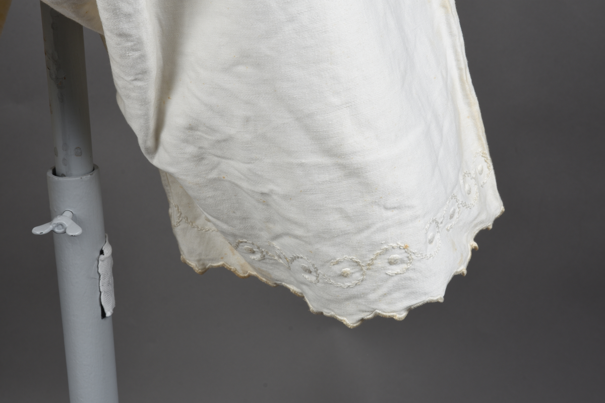 Kombinasjon av bomullslerret. Kraftig kypervevd lerret (twill). Overdelen har brede skulderstropper og er figursydd til en linning i taillen. Benklærne er sydd til samme linning og de har lavt skritt og er helt åpne nedentil. Knappet foran med elleve små knapper. Broderi og tungekant nederst - “løpende hund”-ranke rundt store prikker, tungekant på beina. Noen rustflekker. Lite hull revet bak.