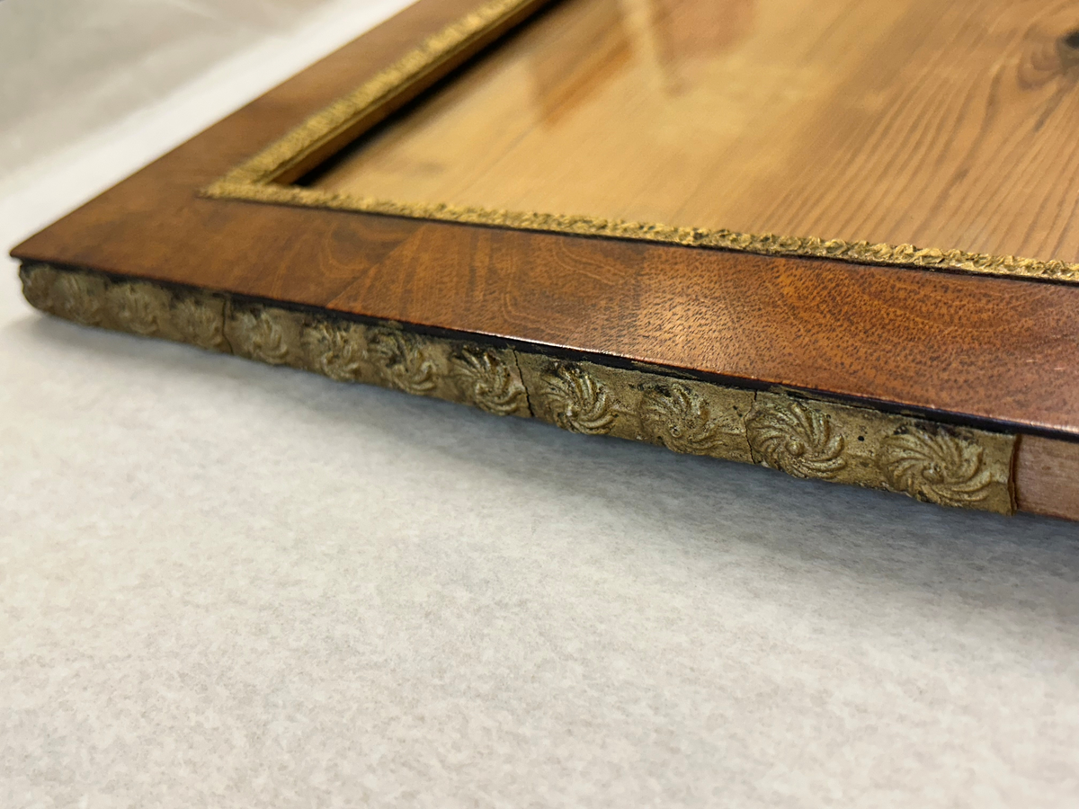 Ram av mahogny med förgylld palmettlist i pastellage med tillhörande glas. Runt ramen löper en förgylld kant med blomrosetter i pastellage. Baksidan av ramen är furu.