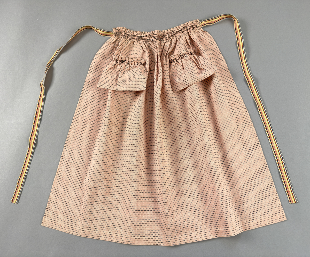 Rosa förkläde, handvävt i tuskaft med inslag av bruna "hopprosor" i bomullgarn. Förklädet har två utanpåhängande *fickor med smocksöm i övre kant. Förklädets midjelinje har också smocksöm sydd med svart tråd i 3mm glesa rader. Traskanten upptill är nervikt mot avigsidan och dolt av ett handvävt band i varprips av bomullsgarn. Bandet är fastsytt med smygstygn runt om samt fortsätter som knytband 62 och 64cm ut på var sida. Långsidorna och nederkant har 3mm respektive 17mm bred dubbelvikt fåll, som är fastsydd med smygstygn. 
* Fickdelen (ca 15x23,5cm) lagd räta mot räta på förklädet är fastsytt med en raksöm ca 8cm lång längs ena fickdelens kortsida. Kortsidans traskant, gömd i en 8mm bred dubbelvikt fåll, vikt mot fickans insida är sydd med smygstygn. Den andra kortsidan är fållad på samma sätt innan den sytts med smocksöm i 5mm glesa rader. Därefter är fickdelen vikt (botten) räta mot räta och ihopsydd med raksöm längs långsidorna, men fickans utsida med smocksöm är lagd 10mm ovanför för att dölja den andra kortsidans fåll. Denna del, 10mm, har sytts fast och fållats på insidan med smygstygn. Se bilder 5-11 på fickan.