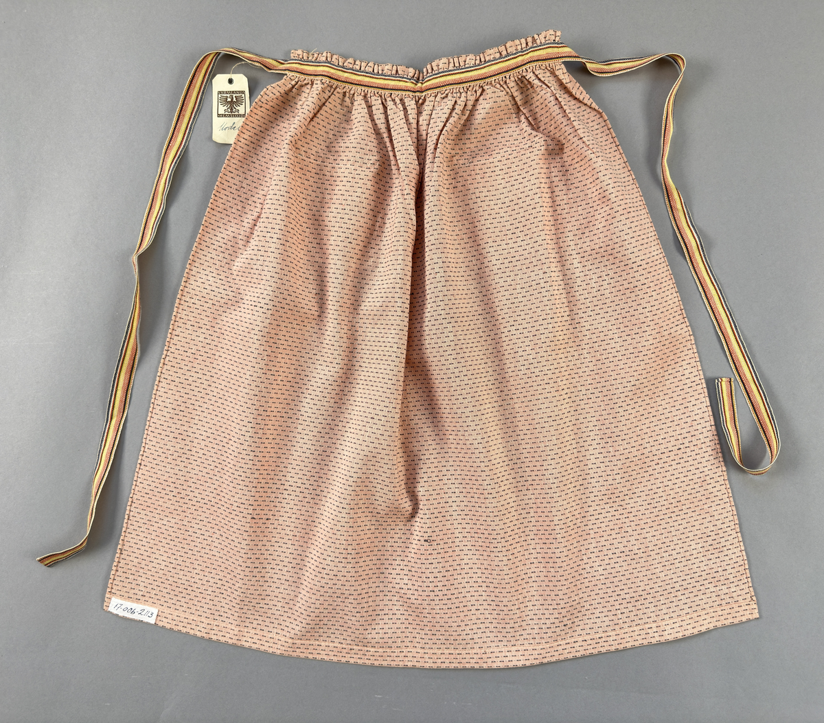 Rosa förkläde, handvävt i tuskaft med inslag av bruna "hopprosor" i bomullgarn. Förklädet har två utanpåhängande *fickor med smocksöm i övre kant. Förklädets midjelinje har också smocksöm sydd med svart tråd i 3mm glesa rader. Traskanten upptill är nervikt mot avigsidan och dolt av ett handvävt band i varprips av bomullsgarn. Bandet är fastsytt med smygstygn runt om samt fortsätter som knytband 62 och 64cm ut på var sida. Långsidorna och nederkant har 3mm respektive 17mm bred dubbelvikt fåll, som är fastsydd med smygstygn. 
* Fickdelen (ca 15x23,5cm) lagd räta mot räta på förklädet är fastsytt med en raksöm ca 8cm lång längs ena fickdelens kortsida. Kortsidans traskant, gömd i en 8mm bred dubbelvikt fåll, vikt mot fickans insida är sydd med smygstygn. Den andra kortsidan är fållad på samma sätt innan den sytts med smocksöm i 5mm glesa rader. Därefter är fickdelen vikt (botten) räta mot räta och ihopsydd med raksöm längs långsidorna, men fickans utsida med smocksöm är lagd 10mm ovanför för att dölja den andra kortsidans fåll. Denna del, 10mm, har sytts fast och fållats på insidan med smygstygn. Se bilder 5-11 på fickan.