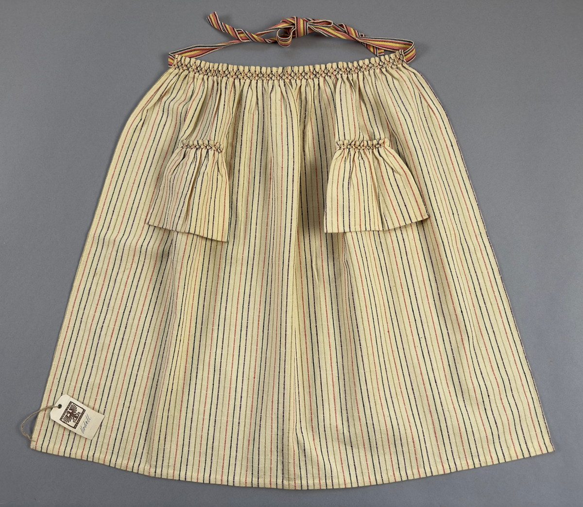 Smalrandigt förkläde, handvävt i tuskaft med inslag av dubbelt garn, som effekt, i lila, grönt, orange samt olivgrönt av bomullgarn. Förklädet har två utanpåhängande *fickor med smocksöm i övre kant. Förklädets midjelinje har också smocksöm sydd med lila tråd i 5mm glesa rader. Traskanten upptill är nervikt mot avigsidan och dolt av ett handvävt band i varprips av bomullsgarn. Bandet är fastsytt med smygstygn runt om samt fortsätter som knytband 55cm ut på var sida. Långsidorna och nederkant har 3mm respektive 10mm bred dubbelvikt fåll, som är fastsydd med smygstygn. 
* Fickdelen (ca 17x27cm) lagd räta mot räta på förklädet är fastsytt med en raksöm ca 8cm lång längs ena fickdelens kortsida. Kortsidans traskant, gömd i en 7mm bred dubbelvikt fåll, vikt mot fickans insida är sydd med smygstygn. Den andra kortsidan är fållad på samma sätt innan den sytts med smocksöm i 5mm glesa rader. Därefter är fickdelen vikt (botten) räta mot räta och ihopsydd med raksöm längs långsidorna, men fickans utsida med smocksöm är lagd 15mm ovanför för att dölja den andra kortsidans fåll. Denna del, 15mm, har sytts fast och fållats på insidan med smygstygn. Se bilder 3-10 på fickan.