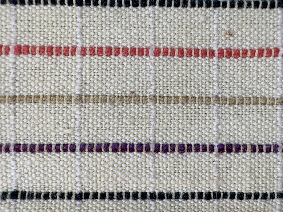 Smalrandigt förkläde, handvävt i tuskaft med inslag av dubbelt garn, som effekt, i lila, grönt, orange samt olivgrönt av bomullgarn. Förklädet har två utanpåhängande *fickor med smocksöm i övre kant. Förklädets midjelinje har också smocksöm sydd med lila tråd i 5mm glesa rader. Traskanten upptill är nervikt mot avigsidan och dolt av ett handvävt band i varprips av bomullsgarn. Bandet är fastsytt med smygstygn runt om samt fortsätter som knytband 55cm ut på var sida. Långsidorna och nederkant har 3mm respektive 10mm bred dubbelvikt fåll, som är fastsydd med smygstygn. 
* Fickdelen (ca 17x27cm) lagd räta mot räta på förklädet är fastsytt med en raksöm ca 8cm lång längs ena fickdelens kortsida. Kortsidans traskant, gömd i en 7mm bred dubbelvikt fåll, vikt mot fickans insida är sydd med smygstygn. Den andra kortsidan är fållad på samma sätt innan den sytts med smocksöm i 5mm glesa rader. Därefter är fickdelen vikt (botten) räta mot räta och ihopsydd med raksöm längs långsidorna, men fickans utsida med smocksöm är lagd 15mm ovanför för att dölja den andra kortsidans fåll. Denna del, 15mm, har sytts fast och fållats på insidan med smygstygn. Se bilder 3-10 på fickan.