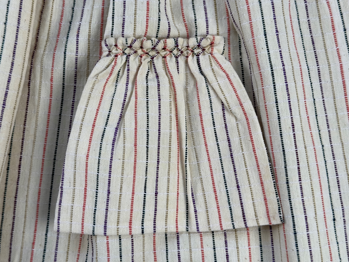 Smalrandigt förkläde, handvävt i tuskaft med inslag av dubbelt garn, som effekt, i lila, grönt, orange samt olivgrönt av bomullgarn. Förklädet har två utanpåhängande *fickor med smocksöm i övre kant. Förklädets midjelinje har också smocksöm sydd med lila tråd i 5mm glesa rader. Traskanten upptill är nervikt mot avigsidan och dolt av ett handvävt band i varprips av bomullsgarn. Bandet är fastsytt med smygstygn runt om samt fortsätter som knytband 55cm ut på var sida. Långsidorna och nederkant har 3mm respektive 10mm bred dubbelvikt fåll, som är fastsydd med smygstygn. 
* Fickdelen (ca 17x27cm) lagd räta mot räta på förklädet är fastsytt med en raksöm ca 8cm lång längs ena fickdelens kortsida. Kortsidans traskant, gömd i en 7mm bred dubbelvikt fåll, vikt mot fickans insida är sydd med smygstygn. Den andra kortsidan är fållad på samma sätt innan den sytts med smocksöm i 5mm glesa rader. Därefter är fickdelen vikt (botten) räta mot räta och ihopsydd med raksöm längs långsidorna, men fickans utsida med smocksöm är lagd 15mm ovanför för att dölja den andra kortsidans fåll. Denna del, 15mm, har sytts fast och fållats på insidan med smygstygn. Se bilder 3-10 på fickan.