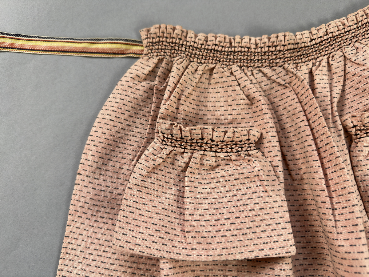 Rosa förkläde, handvävt i tuskaft med inslag av bruna "hopprosor" i bomullgarn. Förklädet har två utanpåhängande *fickor med smocksöm i övre kant. Förklädets midjelinje har också smocksöm sydd med svart tråd i 3mm glesa rader. Traskanten upptill är nervikt mot avigsidan och dolt av ett handvävt band i varprips av bomullsgarn. Bandet är fastsytt med smygstygn runt om samt fortsätter som knytband 62 och 64cm ut på var sida. Långsidorna och nederkant har 3mm respektive 17mm bred dubbelvikt fåll, som är fastsydd med smygstygn. 
* Fickdelen (ca 15x23,5cm) lagd räta mot räta på förklädet är fastsytt med en raksöm ca 8cm lång längs ena fickdelens kortsida. Kortsidans traskant, gömd i en 8mm bred dubbelvikt fåll, vikt mot fickans insida är sydd med smygstygn. Den andra kortsidan är fållad på samma sätt innan den sytts med smocksöm i 5mm glesa rader. Därefter är fickdelen vikt (botten) räta mot räta och ihopsydd med raksöm längs långsidorna, men fickans utsida med smocksöm är lagd 10mm ovanför för att dölja den andra kortsidans fåll. Denna del, 10mm, har sytts fast och fållats på insidan med smygstygn. Se bilder 5-11 på fickan.