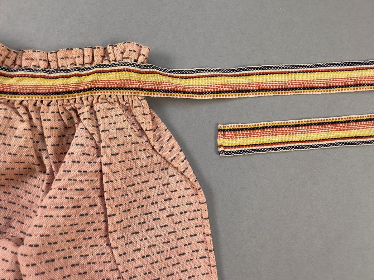 Rosa förkläde, handvävt i tuskaft med inslag av bruna "hopprosor" i bomullgarn. Förklädet har två utanpåhängande *fickor med smocksöm i övre kant. Förklädets midjelinje har också smocksöm sydd med svart tråd i 3mm glesa rader. Traskanten upptill är nervikt mot avigsidan och dolt av ett handvävt band i varprips av bomullsgarn. Bandet är fastsytt med smygstygn runt om samt fortsätter som knytband 62 och 64cm ut på var sida. Långsidorna och nederkant har 3mm respektive 17mm bred dubbelvikt fåll, som är fastsydd med smygstygn. 
* Fickdelen (ca 15x23,5cm) lagd räta mot räta på förklädet är fastsytt med en raksöm ca 8cm lång längs ena fickdelens kortsida. Kortsidans traskant, gömd i en 8mm bred dubbelvikt fåll, vikt mot fickans insida är sydd med smygstygn. Den andra kortsidan är fållad på samma sätt innan den sytts med smocksöm i 5mm glesa rader. Därefter är fickdelen vikt (botten) räta mot räta och ihopsydd med raksöm längs långsidorna, men fickans utsida med smocksöm är lagd 10mm ovanför för att dölja den andra kortsidans fåll. Denna del, 10mm, har sytts fast och fållats på insidan med smygstygn. Se bilder 5-11 på fickan.