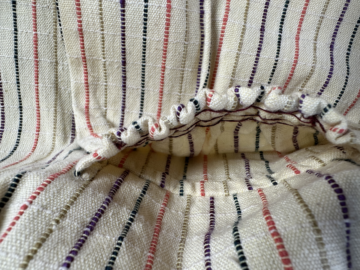 Smalrandigt förkläde, handvävt i tuskaft med inslag av dubbelt garn, som effekt, i lila, grönt, orange samt olivgrönt av bomullgarn. Förklädet har två utanpåhängande *fickor med smocksöm i övre kant. Förklädets midjelinje har också smocksöm sydd med lila tråd i 5mm glesa rader. Traskanten upptill är nervikt mot avigsidan och dolt av ett handvävt band i varprips av bomullsgarn. Bandet är fastsytt med smygstygn runt om samt fortsätter som knytband 55cm ut på var sida. Långsidorna och nederkant har 3mm respektive 10mm bred dubbelvikt fåll, som är fastsydd med smygstygn. 
* Fickdelen (ca 17x27cm) lagd räta mot räta på förklädet är fastsytt med en raksöm ca 8cm lång längs ena fickdelens kortsida. Kortsidans traskant, gömd i en 7mm bred dubbelvikt fåll, vikt mot fickans insida är sydd med smygstygn. Den andra kortsidan är fållad på samma sätt innan den sytts med smocksöm i 5mm glesa rader. Därefter är fickdelen vikt (botten) räta mot räta och ihopsydd med raksöm längs långsidorna, men fickans utsida med smocksöm är lagd 15mm ovanför för att dölja den andra kortsidans fåll. Denna del, 15mm, har sytts fast och fållats på insidan med smygstygn. Se bilder 3-10 på fickan.