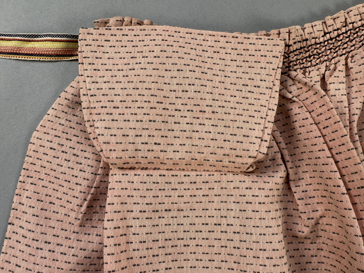 Rosa förkläde, handvävt i tuskaft med inslag av bruna "hopprosor" i bomullgarn. Förklädet har två utanpåhängande *fickor med smocksöm i övre kant. Förklädets midjelinje har också smocksöm sydd med svart tråd i 3mm glesa rader. Traskanten upptill är nervikt mot avigsidan och dolt av ett handvävt band i varprips av bomullsgarn. Bandet är fastsytt med smygstygn runt om samt fortsätter som knytband 62 och 64cm ut på var sida. Långsidorna och nederkant har 3mm respektive 17mm bred dubbelvikt fåll, som är fastsydd med smygstygn. 
* Fickdelen (ca 15x23,5cm) lagd räta mot räta på förklädet är fastsytt med en raksöm ca 8cm lång längs ena fickdelens kortsida. Kortsidans traskant, gömd i en 8mm bred dubbelvikt fåll, vikt mot fickans insida är sydd med smygstygn. Den andra kortsidan är fållad på samma sätt innan den sytts med smocksöm i 5mm glesa rader. Därefter är fickdelen vikt (botten) räta mot räta och ihopsydd med raksöm längs långsidorna, men fickans utsida med smocksöm är lagd 10mm ovanför för att dölja den andra kortsidans fåll. Denna del, 10mm, har sytts fast och fållats på insidan med smygstygn. Se bilder 5-11 på fickan.