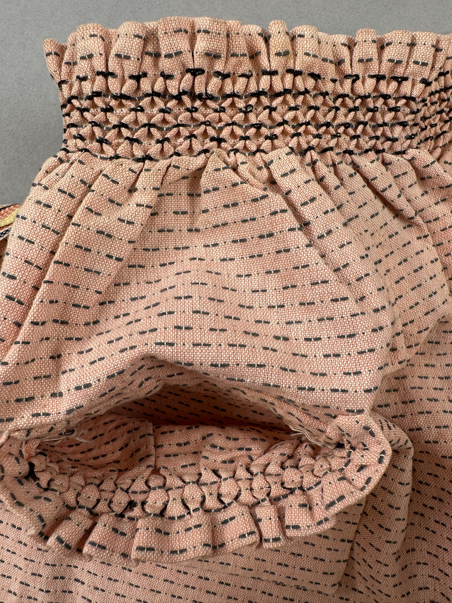 Rosa förkläde, handvävt i tuskaft med inslag av bruna "hopprosor" i bomullgarn. Förklädet har två utanpåhängande *fickor med smocksöm i övre kant. Förklädets midjelinje har också smocksöm sydd med svart tråd i 3mm glesa rader. Traskanten upptill är nervikt mot avigsidan och dolt av ett handvävt band i varprips av bomullsgarn. Bandet är fastsytt med smygstygn runt om samt fortsätter som knytband 62 och 64cm ut på var sida. Långsidorna och nederkant har 3mm respektive 17mm bred dubbelvikt fåll, som är fastsydd med smygstygn. 
* Fickdelen (ca 15x23,5cm) lagd räta mot räta på förklädet är fastsytt med en raksöm ca 8cm lång längs ena fickdelens kortsida. Kortsidans traskant, gömd i en 8mm bred dubbelvikt fåll, vikt mot fickans insida är sydd med smygstygn. Den andra kortsidan är fållad på samma sätt innan den sytts med smocksöm i 5mm glesa rader. Därefter är fickdelen vikt (botten) räta mot räta och ihopsydd med raksöm längs långsidorna, men fickans utsida med smocksöm är lagd 10mm ovanför för att dölja den andra kortsidans fåll. Denna del, 10mm, har sytts fast och fållats på insidan med smygstygn. Se bilder 5-11 på fickan.