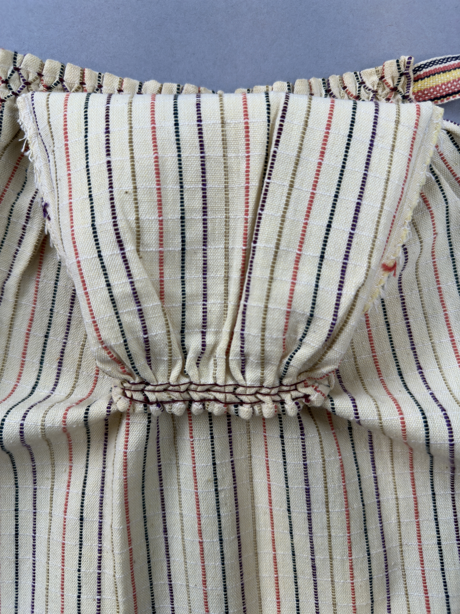 Smalrandigt förkläde, handvävt i tuskaft med inslag av dubbelt garn, som effekt, i lila, grönt, orange samt olivgrönt av bomullgarn. Förklädet har två utanpåhängande *fickor med smocksöm i övre kant. Förklädets midjelinje har också smocksöm sydd med lila tråd i 5mm glesa rader. Traskanten upptill är nervikt mot avigsidan och dolt av ett handvävt band i varprips av bomullsgarn. Bandet är fastsytt med smygstygn runt om samt fortsätter som knytband 55cm ut på var sida. Långsidorna och nederkant har 3mm respektive 10mm bred dubbelvikt fåll, som är fastsydd med smygstygn. 
* Fickdelen (ca 17x27cm) lagd räta mot räta på förklädet är fastsytt med en raksöm ca 8cm lång längs ena fickdelens kortsida. Kortsidans traskant, gömd i en 7mm bred dubbelvikt fåll, vikt mot fickans insida är sydd med smygstygn. Den andra kortsidan är fållad på samma sätt innan den sytts med smocksöm i 5mm glesa rader. Därefter är fickdelen vikt (botten) räta mot räta och ihopsydd med raksöm längs långsidorna, men fickans utsida med smocksöm är lagd 15mm ovanför för att dölja den andra kortsidans fåll. Denna del, 15mm, har sytts fast och fållats på insidan med smygstygn. Se bilder 3-10 på fickan.