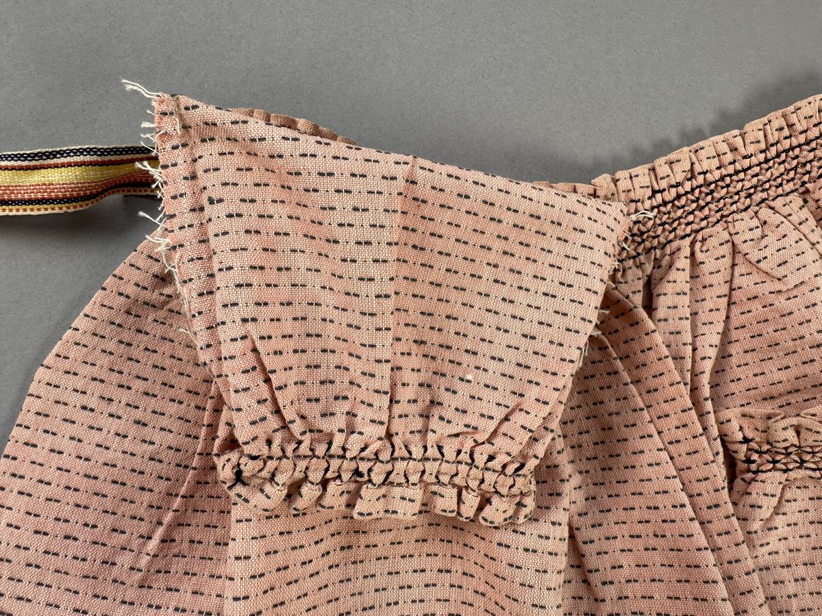 Rosa förkläde, handvävt i tuskaft med inslag av bruna "hopprosor" i bomullgarn. Förklädet har två utanpåhängande *fickor med smocksöm i övre kant. Förklädets midjelinje har också smocksöm sydd med svart tråd i 3mm glesa rader. Traskanten upptill är nervikt mot avigsidan och dolt av ett handvävt band i varprips av bomullsgarn. Bandet är fastsytt med smygstygn runt om samt fortsätter som knytband 62 och 64cm ut på var sida. Långsidorna och nederkant har 3mm respektive 17mm bred dubbelvikt fåll, som är fastsydd med smygstygn. 
* Fickdelen (ca 15x23,5cm) lagd räta mot räta på förklädet är fastsytt med en raksöm ca 8cm lång längs ena fickdelens kortsida. Kortsidans traskant, gömd i en 8mm bred dubbelvikt fåll, vikt mot fickans insida är sydd med smygstygn. Den andra kortsidan är fållad på samma sätt innan den sytts med smocksöm i 5mm glesa rader. Därefter är fickdelen vikt (botten) räta mot räta och ihopsydd med raksöm längs långsidorna, men fickans utsida med smocksöm är lagd 10mm ovanför för att dölja den andra kortsidans fåll. Denna del, 10mm, har sytts fast och fållats på insidan med smygstygn. Se bilder 5-11 på fickan.