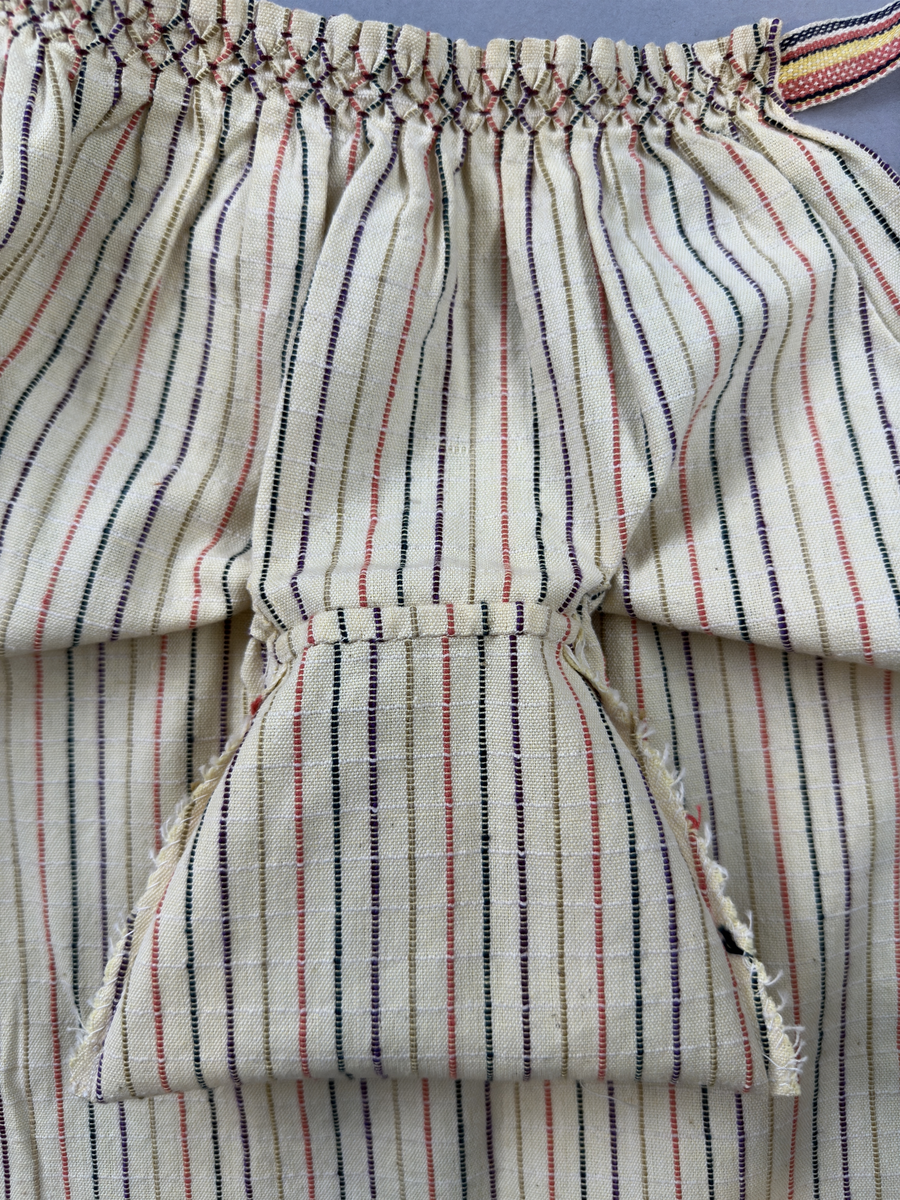 Smalrandigt förkläde, handvävt i tuskaft med inslag av dubbelt garn, som effekt, i lila, grönt, orange samt olivgrönt av bomullgarn. Förklädet har två utanpåhängande *fickor med smocksöm i övre kant. Förklädets midjelinje har också smocksöm sydd med lila tråd i 5mm glesa rader. Traskanten upptill är nervikt mot avigsidan och dolt av ett handvävt band i varprips av bomullsgarn. Bandet är fastsytt med smygstygn runt om samt fortsätter som knytband 55cm ut på var sida. Långsidorna och nederkant har 3mm respektive 10mm bred dubbelvikt fåll, som är fastsydd med smygstygn. 
* Fickdelen (ca 17x27cm) lagd räta mot räta på förklädet är fastsytt med en raksöm ca 8cm lång längs ena fickdelens kortsida. Kortsidans traskant, gömd i en 7mm bred dubbelvikt fåll, vikt mot fickans insida är sydd med smygstygn. Den andra kortsidan är fållad på samma sätt innan den sytts med smocksöm i 5mm glesa rader. Därefter är fickdelen vikt (botten) räta mot räta och ihopsydd med raksöm längs långsidorna, men fickans utsida med smocksöm är lagd 15mm ovanför för att dölja den andra kortsidans fåll. Denna del, 15mm, har sytts fast och fållats på insidan med smygstygn. Se bilder 3-10 på fickan.