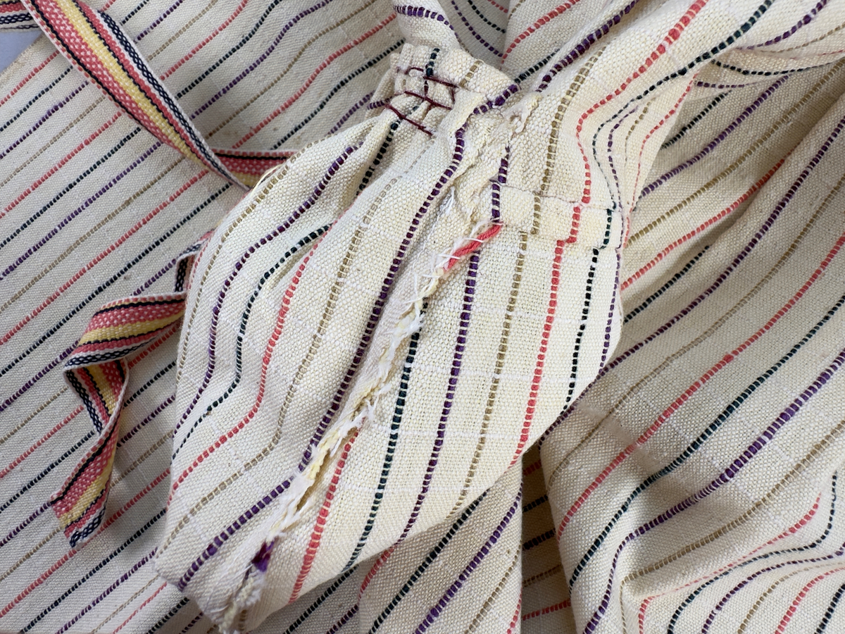 Smalrandigt förkläde, handvävt i tuskaft med inslag av dubbelt garn, som effekt, i lila, grönt, orange samt olivgrönt av bomullgarn. Förklädet har två utanpåhängande *fickor med smocksöm i övre kant. Förklädets midjelinje har också smocksöm sydd med lila tråd i 5mm glesa rader. Traskanten upptill är nervikt mot avigsidan och dolt av ett handvävt band i varprips av bomullsgarn. Bandet är fastsytt med smygstygn runt om samt fortsätter som knytband 55cm ut på var sida. Långsidorna och nederkant har 3mm respektive 10mm bred dubbelvikt fåll, som är fastsydd med smygstygn. 
* Fickdelen (ca 17x27cm) lagd räta mot räta på förklädet är fastsytt med en raksöm ca 8cm lång längs ena fickdelens kortsida. Kortsidans traskant, gömd i en 7mm bred dubbelvikt fåll, vikt mot fickans insida är sydd med smygstygn. Den andra kortsidan är fållad på samma sätt innan den sytts med smocksöm i 5mm glesa rader. Därefter är fickdelen vikt (botten) räta mot räta och ihopsydd med raksöm längs långsidorna, men fickans utsida med smocksöm är lagd 15mm ovanför för att dölja den andra kortsidans fåll. Denna del, 15mm, har sytts fast och fållats på insidan med smygstygn. Se bilder 3-10 på fickan.