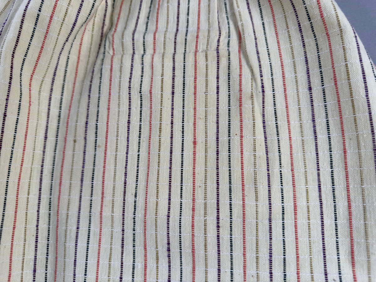 Smalrandigt förkläde, handvävt i tuskaft med inslag av dubbelt garn, som effekt, i lila, grönt, orange samt olivgrönt av bomullgarn. Förklädet har två utanpåhängande *fickor med smocksöm i övre kant. Förklädets midjelinje har också smocksöm sydd med lila tråd i 5mm glesa rader. Traskanten upptill är nervikt mot avigsidan och dolt av ett handvävt band i varprips av bomullsgarn. Bandet är fastsytt med smygstygn runt om samt fortsätter som knytband 55cm ut på var sida. Långsidorna och nederkant har 3mm respektive 10mm bred dubbelvikt fåll, som är fastsydd med smygstygn. 
* Fickdelen (ca 17x27cm) lagd räta mot räta på förklädet är fastsytt med en raksöm ca 8cm lång längs ena fickdelens kortsida. Kortsidans traskant, gömd i en 7mm bred dubbelvikt fåll, vikt mot fickans insida är sydd med smygstygn. Den andra kortsidan är fållad på samma sätt innan den sytts med smocksöm i 5mm glesa rader. Därefter är fickdelen vikt (botten) räta mot räta och ihopsydd med raksöm längs långsidorna, men fickans utsida med smocksöm är lagd 15mm ovanför för att dölja den andra kortsidans fåll. Denna del, 15mm, har sytts fast och fållats på insidan med smygstygn. Se bilder 3-10 på fickan.