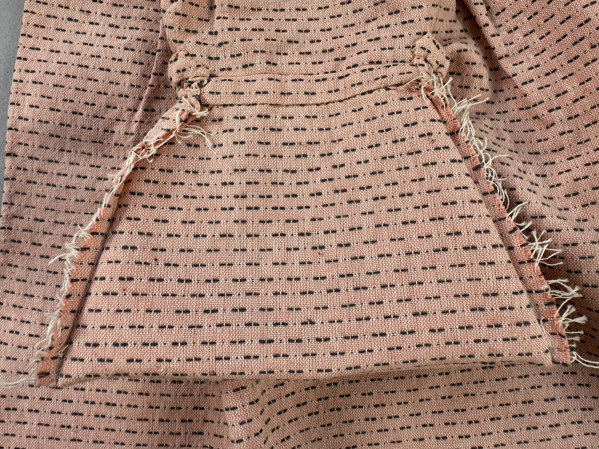 Rosa förkläde, handvävt i tuskaft med inslag av bruna "hopprosor" i bomullgarn. Förklädet har två utanpåhängande *fickor med smocksöm i övre kant. Förklädets midjelinje har också smocksöm sydd med svart tråd i 3mm glesa rader. Traskanten upptill är nervikt mot avigsidan och dolt av ett handvävt band i varprips av bomullsgarn. Bandet är fastsytt med smygstygn runt om samt fortsätter som knytband 62 och 64cm ut på var sida. Långsidorna och nederkant har 3mm respektive 17mm bred dubbelvikt fåll, som är fastsydd med smygstygn. 
* Fickdelen (ca 15x23,5cm) lagd räta mot räta på förklädet är fastsytt med en raksöm ca 8cm lång längs ena fickdelens kortsida. Kortsidans traskant, gömd i en 8mm bred dubbelvikt fåll, vikt mot fickans insida är sydd med smygstygn. Den andra kortsidan är fållad på samma sätt innan den sytts med smocksöm i 5mm glesa rader. Därefter är fickdelen vikt (botten) räta mot räta och ihopsydd med raksöm längs långsidorna, men fickans utsida med smocksöm är lagd 10mm ovanför för att dölja den andra kortsidans fåll. Denna del, 10mm, har sytts fast och fållats på insidan med smygstygn. Se bilder 5-11 på fickan.