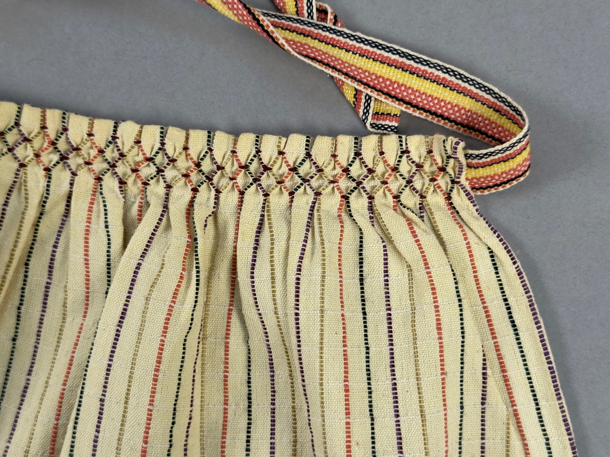 Smalrandigt förkläde, handvävt i tuskaft med inslag av dubbelt garn, som effekt, i lila, grönt, orange samt olivgrönt av bomullgarn. Förklädet har två utanpåhängande *fickor med smocksöm i övre kant. Förklädets midjelinje har också smocksöm sydd med lila tråd i 5mm glesa rader. Traskanten upptill är nervikt mot avigsidan och dolt av ett handvävt band i varprips av bomullsgarn. Bandet är fastsytt med smygstygn runt om samt fortsätter som knytband 55cm ut på var sida. Långsidorna och nederkant har 3mm respektive 10mm bred dubbelvikt fåll, som är fastsydd med smygstygn. 
* Fickdelen (ca 17x27cm) lagd räta mot räta på förklädet är fastsytt med en raksöm ca 8cm lång längs ena fickdelens kortsida. Kortsidans traskant, gömd i en 7mm bred dubbelvikt fåll, vikt mot fickans insida är sydd med smygstygn. Den andra kortsidan är fållad på samma sätt innan den sytts med smocksöm i 5mm glesa rader. Därefter är fickdelen vikt (botten) räta mot räta och ihopsydd med raksöm längs långsidorna, men fickans utsida med smocksöm är lagd 15mm ovanför för att dölja den andra kortsidans fåll. Denna del, 15mm, har sytts fast och fållats på insidan med smygstygn. Se bilder 3-10 på fickan.