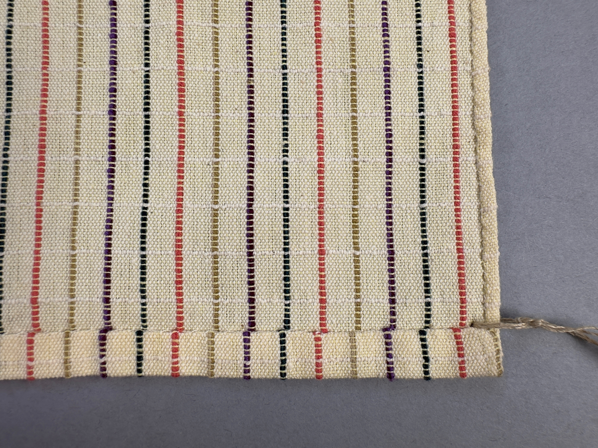 Smalrandigt förkläde, handvävt i tuskaft med inslag av dubbelt garn, som effekt, i lila, grönt, orange samt olivgrönt av bomullgarn. Förklädet har två utanpåhängande *fickor med smocksöm i övre kant. Förklädets midjelinje har också smocksöm sydd med lila tråd i 5mm glesa rader. Traskanten upptill är nervikt mot avigsidan och dolt av ett handvävt band i varprips av bomullsgarn. Bandet är fastsytt med smygstygn runt om samt fortsätter som knytband 55cm ut på var sida. Långsidorna och nederkant har 3mm respektive 10mm bred dubbelvikt fåll, som är fastsydd med smygstygn. 
* Fickdelen (ca 17x27cm) lagd räta mot räta på förklädet är fastsytt med en raksöm ca 8cm lång längs ena fickdelens kortsida. Kortsidans traskant, gömd i en 7mm bred dubbelvikt fåll, vikt mot fickans insida är sydd med smygstygn. Den andra kortsidan är fållad på samma sätt innan den sytts med smocksöm i 5mm glesa rader. Därefter är fickdelen vikt (botten) räta mot räta och ihopsydd med raksöm längs långsidorna, men fickans utsida med smocksöm är lagd 15mm ovanför för att dölja den andra kortsidans fåll. Denna del, 15mm, har sytts fast och fållats på insidan med smygstygn. Se bilder 3-10 på fickan.