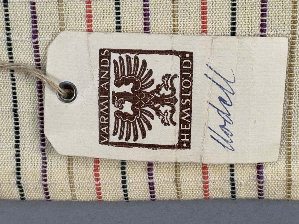 Smalrandigt förkläde, handvävt i tuskaft med inslag av dubbelt garn, som effekt, i lila, grönt, orange samt olivgrönt av bomullgarn. Förklädet har två utanpåhängande *fickor med smocksöm i övre kant. Förklädets midjelinje har också smocksöm sydd med lila tråd i 5mm glesa rader. Traskanten upptill är nervikt mot avigsidan och dolt av ett handvävt band i varprips av bomullsgarn. Bandet är fastsytt med smygstygn runt om samt fortsätter som knytband 55cm ut på var sida. Långsidorna och nederkant har 3mm respektive 10mm bred dubbelvikt fåll, som är fastsydd med smygstygn. 
* Fickdelen (ca 17x27cm) lagd räta mot räta på förklädet är fastsytt med en raksöm ca 8cm lång längs ena fickdelens kortsida. Kortsidans traskant, gömd i en 7mm bred dubbelvikt fåll, vikt mot fickans insida är sydd med smygstygn. Den andra kortsidan är fållad på samma sätt innan den sytts med smocksöm i 5mm glesa rader. Därefter är fickdelen vikt (botten) räta mot räta och ihopsydd med raksöm längs långsidorna, men fickans utsida med smocksöm är lagd 15mm ovanför för att dölja den andra kortsidans fåll. Denna del, 15mm, har sytts fast och fållats på insidan med smygstygn. Se bilder 3-10 på fickan.