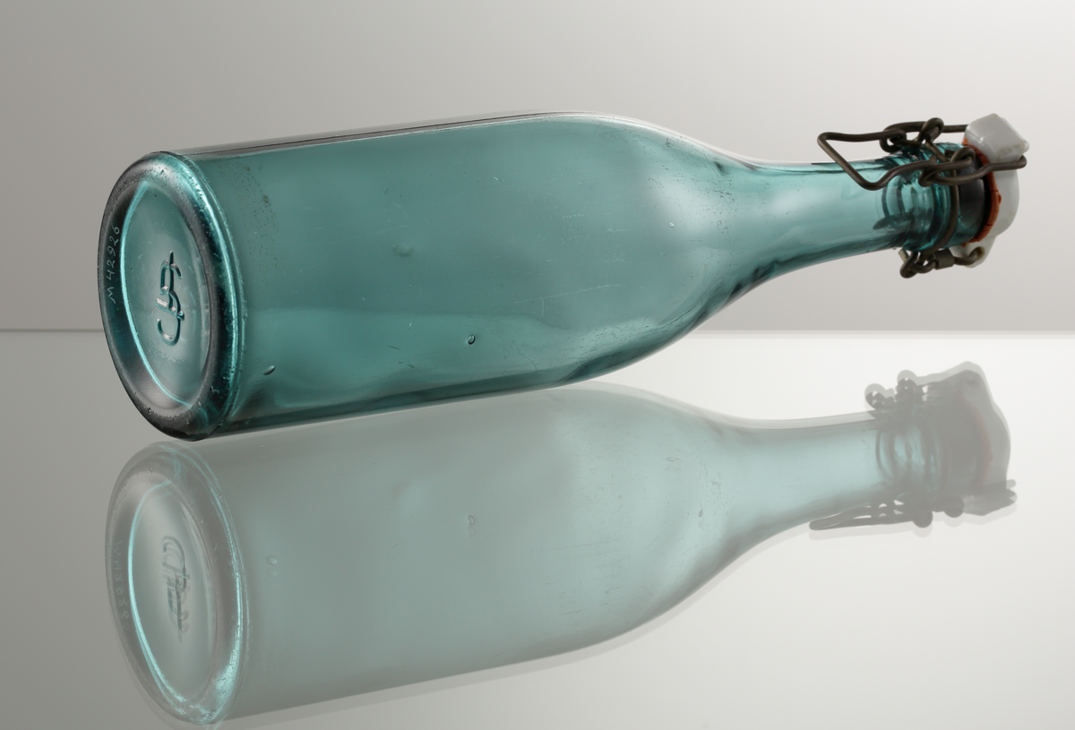 Grön mineralvattenflaska, med patentkork.
Cylindrisk i nedre delen, mjuk övergång i konad hals.
Patentkork (porslin, gummiring, metalltråd.
Relief i botten: "TG" (Trelleborgs glasbruk).
Sodaglas.

Gåva av Ivar Scheutz 1985.