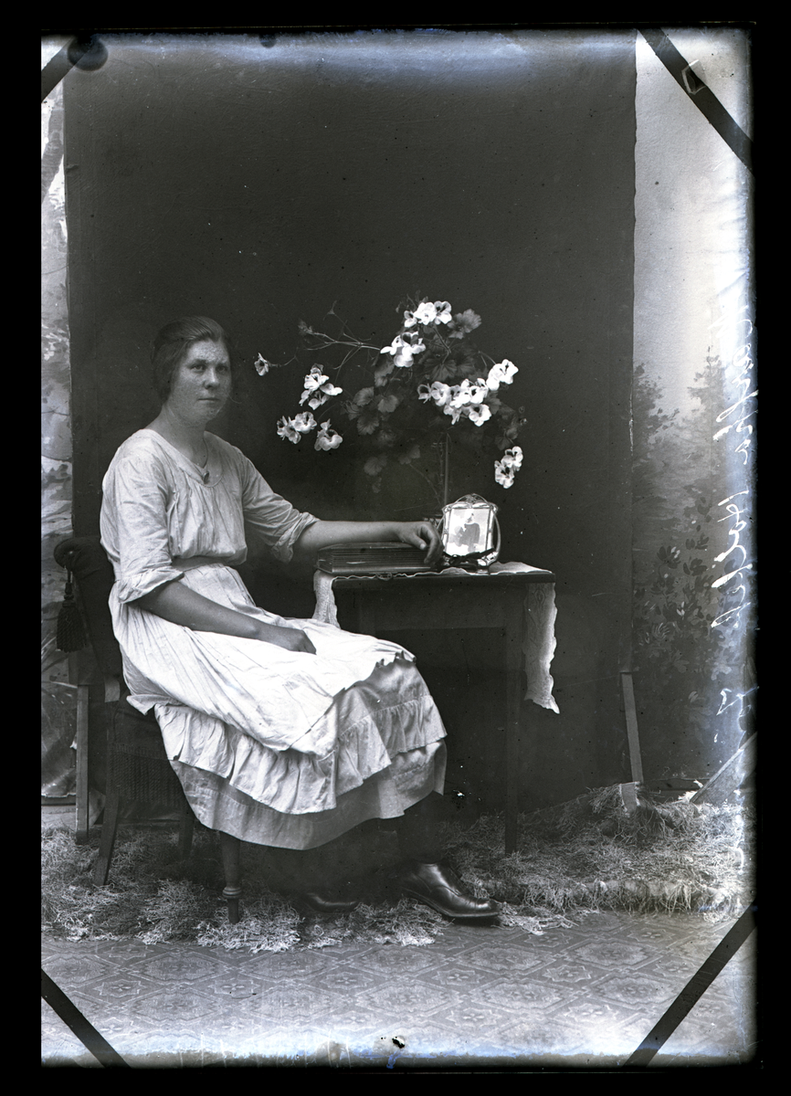 Atelierfoto. En kvinne sitter ved et bord med et bilde og blomster på.
Skrevet på negativet: Martha Holtet Jømna (se bilde GM_MG.01941_1).