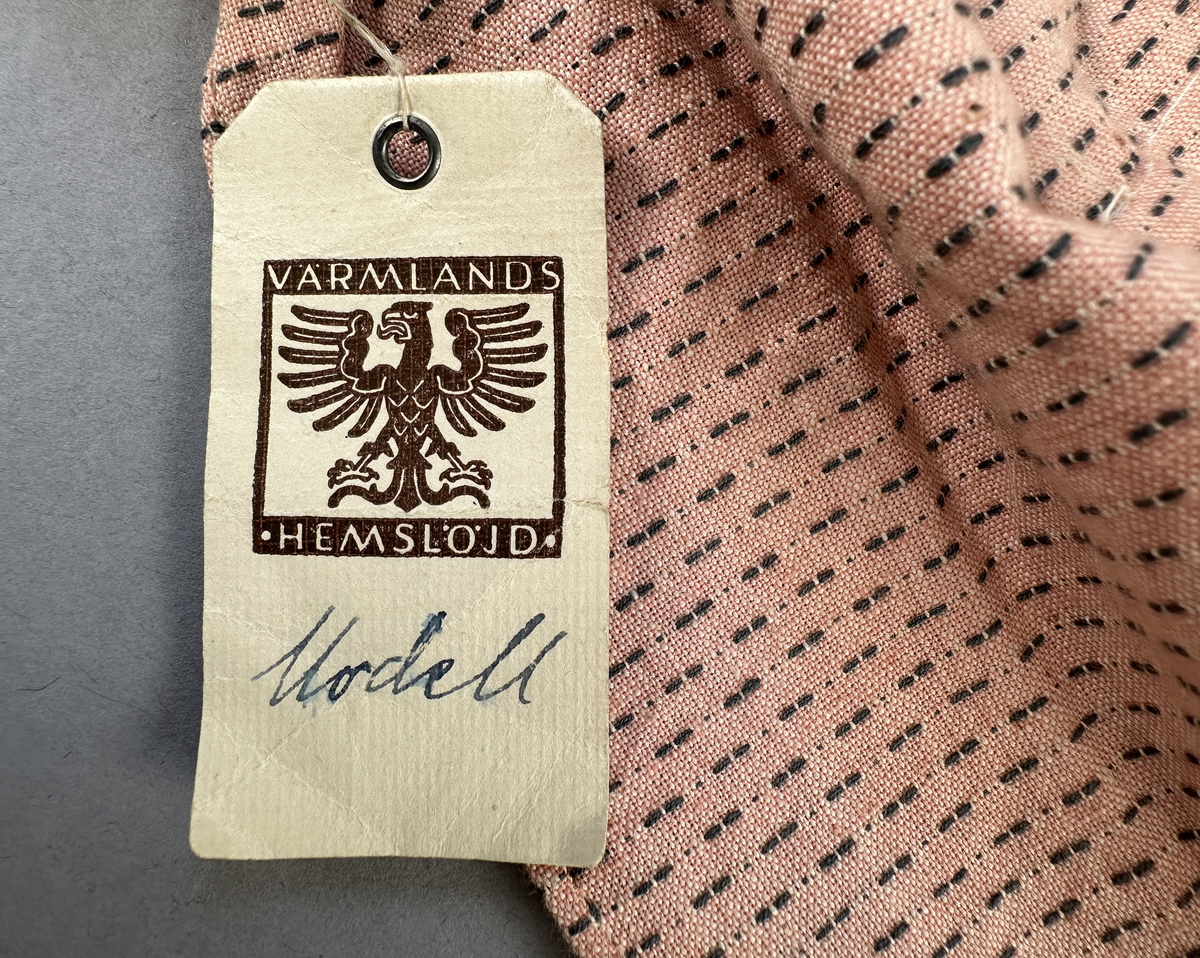 Rosa förkläde, handvävt i tuskaft med inslag av bruna "hopprosor" i bomullgarn. Förklädet har två utanpåhängande *fickor med smocksöm i övre kant. Förklädets midjelinje har också smocksöm sydd med svart tråd i 3mm glesa rader. Traskanten upptill är nervikt mot avigsidan och dolt av ett handvävt band i varprips av bomullsgarn. Bandet är fastsytt med smygstygn runt om samt fortsätter som knytband 62 och 64cm ut på var sida. Långsidorna och nederkant har 3mm respektive 17mm bred dubbelvikt fåll, som är fastsydd med smygstygn. 
* Fickdelen (ca 15x23,5cm) lagd räta mot räta på förklädet är fastsytt med en raksöm ca 8cm lång längs ena fickdelens kortsida. Kortsidans traskant, gömd i en 8mm bred dubbelvikt fåll, vikt mot fickans insida är sydd med smygstygn. Den andra kortsidan är fållad på samma sätt innan den sytts med smocksöm i 5mm glesa rader. Därefter är fickdelen vikt (botten) räta mot räta och ihopsydd med raksöm längs långsidorna, men fickans utsida med smocksöm är lagd 10mm ovanför för att dölja den andra kortsidans fåll. Denna del, 10mm, har sytts fast och fållats på insidan med smygstygn. Se bilder 5-11 på fickan.