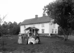 En familj vid ett bord i trädgården framför okänt bostadshus. Ca. 1910.