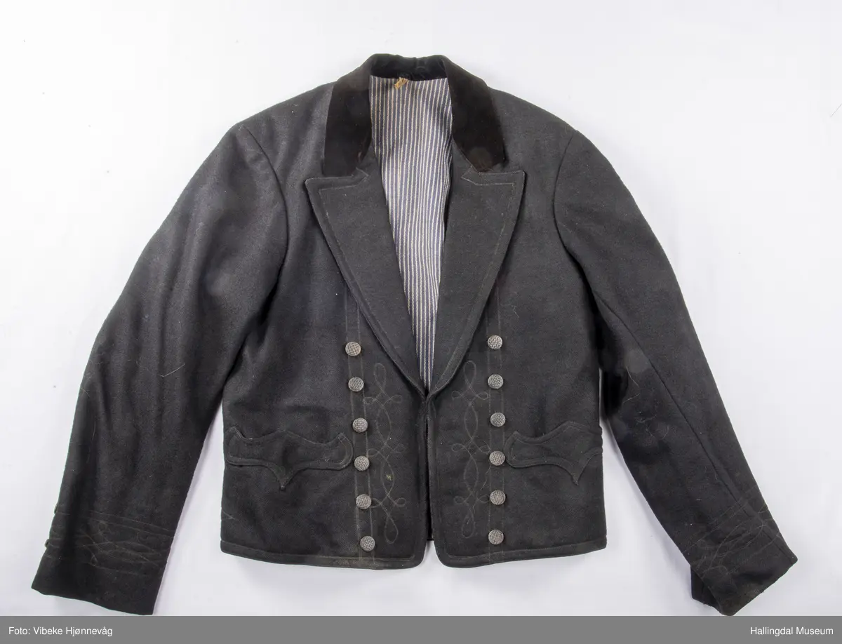 a) åpen mannstrøye, b) mannsvest, c) langbukse, d) halsklut og tilhørende også e) hatt. Open trøye m svarte knappar, dobbelspent vest. Svart m blått, kvitrundut for, rindutt vestebakklut. Sauma maskinsaum til dekor på ermekragar og framside. Ferdig sydd halsklut i silketøy. Filthatt med silkebånd og silkefor.