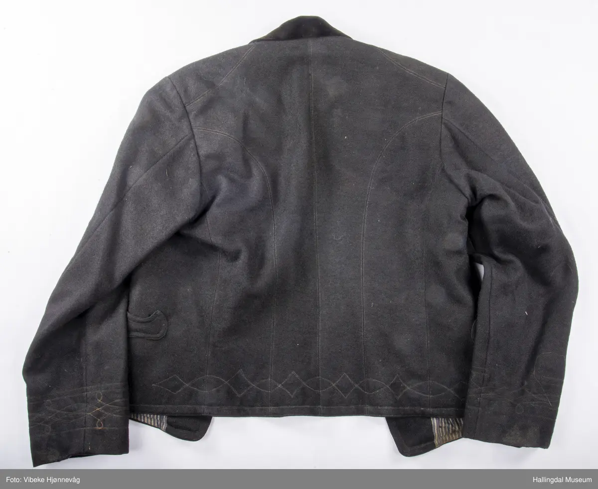 a) åpen mannstrøye, b) mannsvest, c) langbukse, d) halsklut og tilhørende også e) hatt. Open trøye m svarte knappar, dobbelspent vest. Svart m blått, kvitrundut for, rindutt vestebakklut. Sauma maskinsaum til dekor på ermekragar og framside. Ferdig sydd halsklut i silketøy. Filthatt med silkebånd og silkefor.