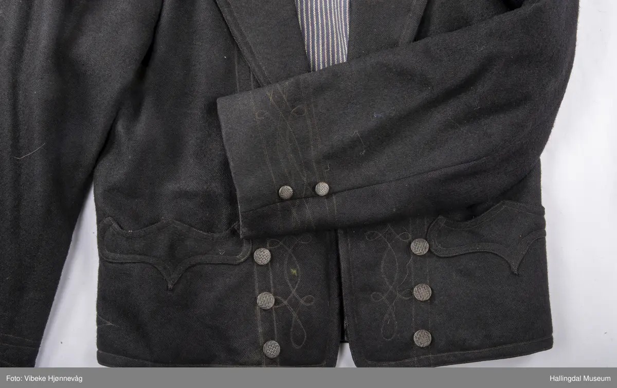 a) åpen mannstrøye, b) mannsvest, c) langbukse, d) halsklut og tilhørende også e) hatt. Open trøye m svarte knappar, dobbelspent vest. Svart m blått, kvitrundut for, rindutt vestebakklut. Sauma maskinsaum til dekor på ermekragar og framside. Ferdig sydd halsklut i silketøy. Filthatt med silkebånd og silkefor.