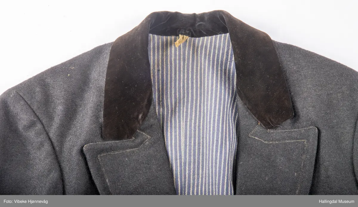 a) åpen mannstrøye, b) mannsvest, c) langbukse, d) halsklut og tilhørende også e) hatt. Open trøye m svarte knappar, dobbelspent vest. Svart m blått, kvitrundut for, rindutt vestebakklut. Sauma maskinsaum til dekor på ermekragar og framside. Ferdig sydd halsklut i silketøy. Filthatt med silkebånd og silkefor.