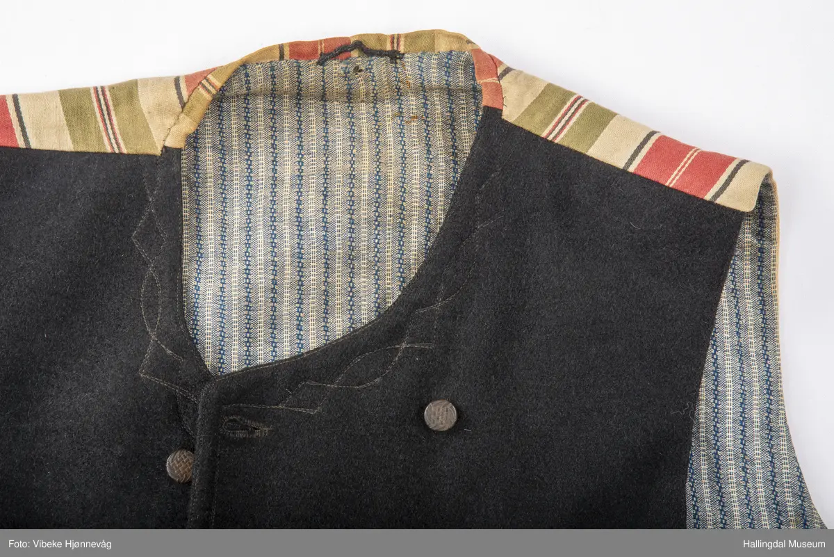a) åpen mannstrøye, b) mannsvest, c) langbukse, d) halsklut og tilhørende også e) hatt. Open trøye m svarte knappar, dobbelspent vest. Svart m blått, kvitrundut for, rindutt vestebakklut. Sauma maskinsaum til dekor på ermekragar og framside. Ferdig sydd halsklut i silketøy. Filthatt med silkebånd og silkefor.