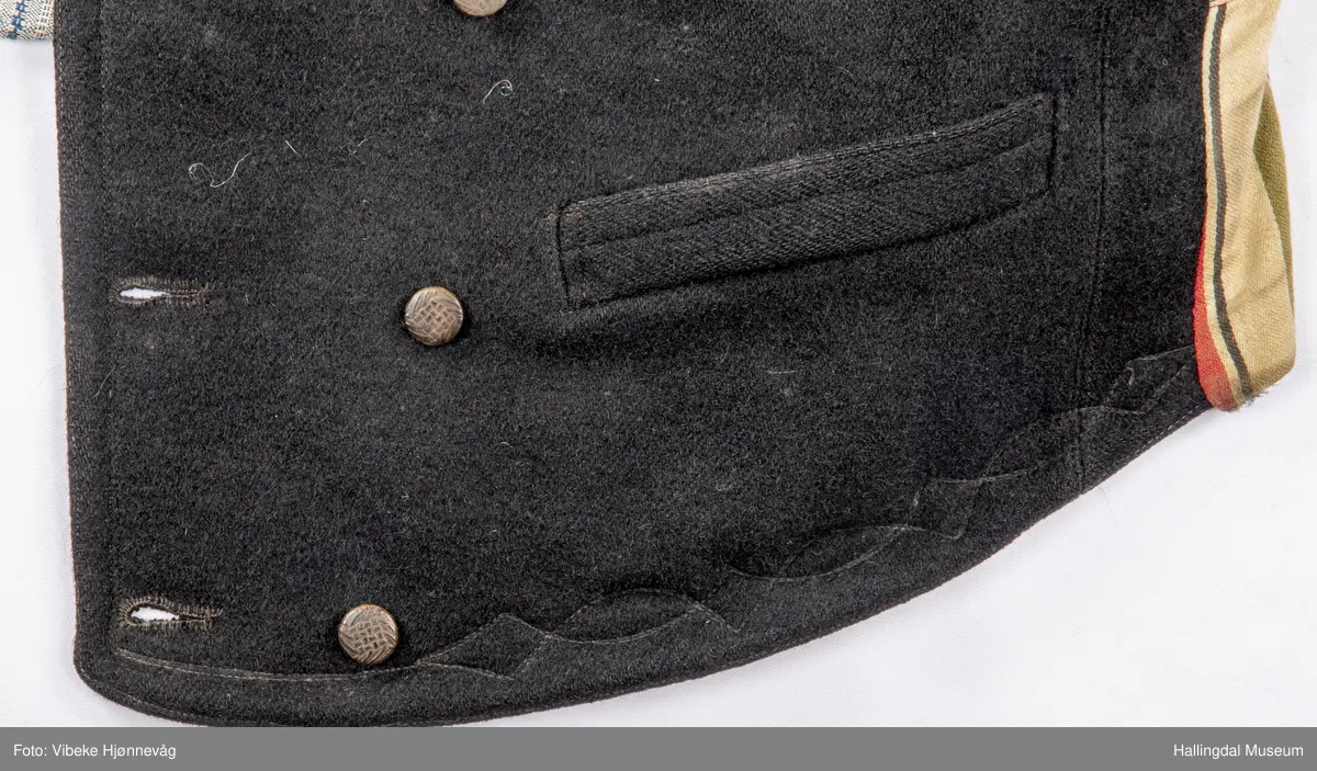 a) åpen mannstrøye, b) mannsvest, c) langbukse, d) halsklut og tilhørende også e) hatt. Open trøye m svarte knappar, dobbelspent vest. Svart m blått, kvitrundut for, rindutt vestebakklut. Sauma maskinsaum til dekor på ermekragar og framside. Ferdig sydd halsklut i silketøy. Filthatt med silkebånd og silkefor.